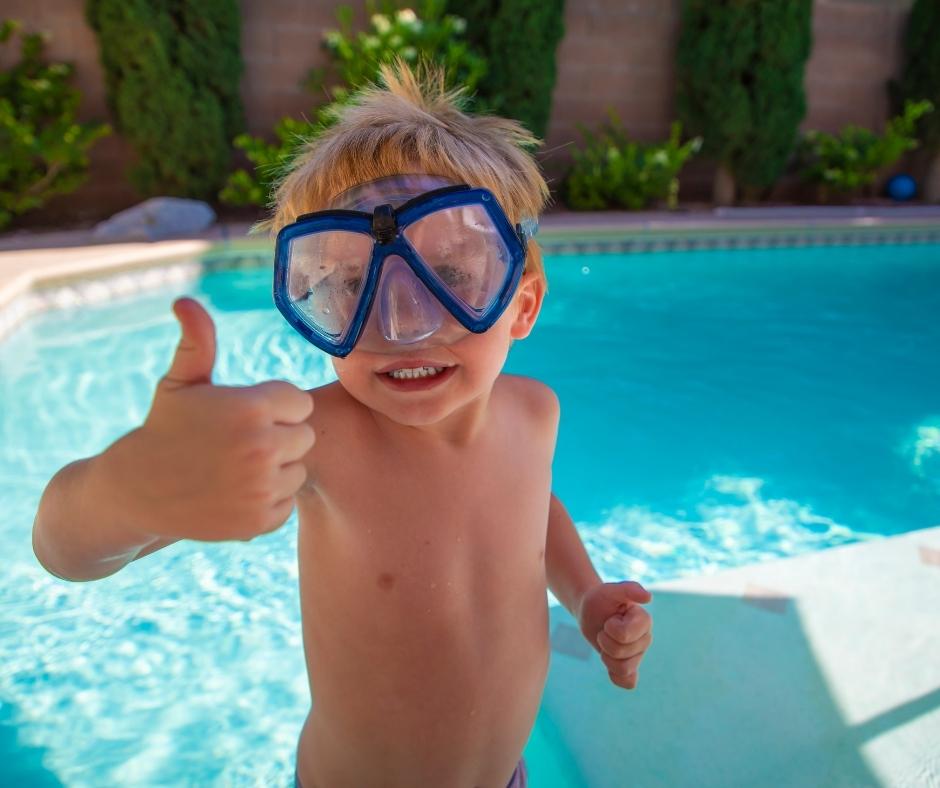 Little person with googles giving a thumbs up with a residential pool behind him
